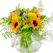 Bursting with life and local colour, this stunning bouquet is anchored by grand, golden sunflowers with plush, chocolatey centers, artfully arranged by a Golders Green florist who knows the neighbourhood's fondness for cheerful, statement blooms. These sunflowers command attention, their petals unfurling like tiny rays of sunlight, while miniature yellow filler flowers-perhaps solidago or mimosa-nestle between them, echoing shades of Hampstead Heath's wildflowers. Subtle notes of soft coral-orange, seen in the petite, cupped accent blooms, introduce a contemporary twist, hinting at creative inspiration drawn from the area's eclectic arts scene. Fern fronds, vivid and feathery, spill outward in all directions, blending seamlessly with touches of blue-green eucalyptus and zesty lime foliage that complete the lively palette. The clear, rounded vase reveals stems bathed in fresh water-an invitation to linger and admire the careful craftsmanship. Bathed in bright, natural light, the arrangement almost hums with positive energy-an ideal display for welcoming in a new season, celebrating a birthday, or bringing a piece of Golders Green's happiness into any home.