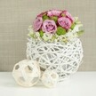 An artfully styled floral centrepiece rests on a bright white tabletop, evoking the calm elegance of a wedding reception set just off Golders Green Road. The arrangement is anchored by a unique round vase made from slender white twigs or rattan strands, woven into an open, globe-like structure so that little windows of negative space show through, lending the design an airy, sculptural quality. Rising from this base is a lush, domed bouquet of roses in delicate shades of pink and lavender, their petals unfurling in soft spirals, with some edges deepening to a richer pink that adds depth and romance. Tucked between these generous blooms are crisp green button chrysanthemums, their compact, bobbly forms bringing a lively texture and a shot of fresh colour that keeps the palette feeling modern rather than overly sweet. Around the foot of the bouquet, tiny white and cream filler flowers cluster together, suggesting baby's breath or other fine-petalled blossoms, creating a gentle cloud that relaxes the edge of the arrangement and lets it appear to spill naturally over the rim. To the left of the main vase, two decorative orbs constructed from pale, natural wicker sit on the same white surface, one larger and one smaller, their interlaced strands echoing the woven container and tying the composition together. Behind, a softly out-of-focus backdrop in warm beige, reminiscent of linen or finely woven fabric, lends subtle texture without competing with the flowers. The overall mood is peaceful, refined, and slightly romantic, ideal for describing a wedding table arrangement or chic home décor piece suited to celebrations in and around Golders Green.