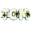 Displayed against a simple white background, this personalised funeral flower tribute spells out the name TOM in large block letters, arranged side by side on a slender, dark supporting rail that gives the impression they are gently floating. Every letter is completely carpeted with compact white chrysanthemums, creating a uniform, cushion-like appearance with subtle shadows between the petals that add depth and softness. A royal-blue satin ribbon follows the outline of each letter, neatly pleated and folded so that its sheen catches the light and provides a strong yet respectful contrast to the all-white centre. At thoughtfully chosen points across the design, the calm surface is broken by rich floral sprays: deep red roses at full or near-full bloom, clusters of small white or cream spray roses, and a few narrow, trumpet-shaped white flowers with the airy look of freesia. These are threaded with fresh green foliage-ferns, slender leaves, and small sprays of greenery-which arcs outwards, bringing a touch of natural looseness to the otherwise structured shape. The colour palette of white, blue, red, and green feels both traditional and heartfelt, ideal for a farewell in the Golders Green area, perhaps on the way to a local cemetery or crematorium. This piece is clearly intended as a bespoke memorial for someone named Tom, crafted with care to offer a visible expression of sorrow, gratitude, and love during a time of grief, allowing family and friends to see his name honoured in flowers.