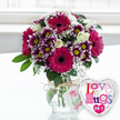 The photograph captures a joyful gift ensemble that could easily be waiting on a kitchen table in Golders Green after a thoughtful surprise delivery. At the heart of the scene is a full, rounded bouquet held in a classic clear glass vase, where smooth sides reveal a criss-cross of green stems resting in clear water. Around the vase neck, a strand of natural jute twine is tied in a simple, rustic bow, from which hangs a slim white tag decorated with a pink rose icon and friendly green lettering that hints at a florist's personal touch. The bouquet itself is a vibrant celebration of purple and pink tones: large magenta gerbera daisies with plush petals and nearly black centres radiate outward, giving the piece its bold personality. Interspersed are numerous smaller chrysanthemums whose deep purple petals fade to snowy white at their tips, framing bright yellow centres that glow with an almost sunshine-like warmth. Creamy white roses, some in bud, others opening, tuck themselves gently among the more vivid blooms, creating layers of softness and elegance. Tiny white flowers of baby's breath scatter through the arrangement like a delicate mist, while fresh green leaves and foliage provide a verdant backdrop that enhances every colour. To the lower right of the vase, overlapping it slightly, a heart-shaped foil balloon shines with a metallic silver border and white face. Across its surface, the playful message Love n' Hugs 24-7 appears in a mosaic of pink, purple, blue, and patterned letters, punctuated with small hearts, reinforcing the mood of constant affection. The softly lit, blurred white background suggests a bright room with a nearby window, allowing the cheerful purples, creams, and greens to stand out as a perfect token of love, comfort, or celebration for someone special in Golders Green.