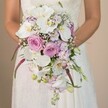 Bathed in gentle daylight within a serene Golders Green backdrop, a bride stands elegantly at ease, her arms gathered around a grand, teardrop-shaped cascading bouquet that shimmers with colour and texture. The vivid arrangement draws immediate attention, composed of wide, pure white Phalaenopsis orchids that stretch gracefully and anchor the composition. These luminous blooms are nestled among an array of soft blush pink and lavender roses, each petal velvety and unique, some unfurling in full glory while others remain tender buds. Whispers of gypsophila-those airy, cloudlike baby's breath sprays-add a sensation of lightness and romance, diffusing between larger blooms. Streaks of fuchsia Amaranthus cascade downwards, their vibrant hue and tactile surface bringing depth and dramatic flow to the bouquet's silhouette. Delicate sprigs of lavender and light purple, hinting at stock or sweet pea, layer further visual intrigue and scent. A trace of green foliage and unopened buds peek from the bouquet, echoing the freshness and expertise of Golders Green's best florists. The bride's lacy, ivory gown, visible from mid-torso, amplifies the sense of tradition amidst a modern design. The soft, monochromatic background-a creamy, muted wall-centres the vibrant flowers, ensuring every detail stands out. Perfect for weddings across Golders Green and NW11, this bouquet is an inspiring blend of artistry, nature, and heartfelt celebration, offering couples an exquisite vision for their special day.