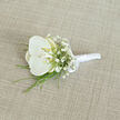 An elegant white orchid boutonnière is shown resting on a softly woven, light beige fabric, as if laid out on a tailor's table before a Golders Green wedding. The central orchid is pristine and luminous, with wide, velvety petals unfolding in a perfect oval, their creamy white surface catching the light with a gentle sheen. At the heart of the bloom, a touch of pale yellow and cream glows from the throat, adding depth and warmth to the otherwise cool palette. Surrounding the orchid, clusters of white baby's breath form a delicate mist of miniature blossoms, each tiny flower contributing to a sense of softness and romance. Slender green blades of foliage extend subtly from the base, their fresh, bright tone introducing a graceful line and a hint of natural movement. The stems of the flowers are carefully aligned and wrapped in smooth white satin ribbon, the fabric tightly wound to form a narrow, tidy handle that would sit comfortably on the lapel of a finely cut suit. A small satin bow is tied near the top of the wrap, its loops neat and proportional, suggesting a florist's practiced touch and attention to finish. The background cloth, with its natural, linen-like weave in shades of light tan, provides an understated backdrop, reminiscent of the calm interiors of local venues off Finchley Road or near Golders Green Station. The light in the image is soft and even, revealing textures without harsh shadow, and the overall mood is one of quiet anticipation, as though this boutonnière is waiting to be pinned on just before the ceremony begins.