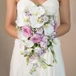 In this radiant portrait set against a softly muted, biscuit-toned background, a bride prepares for her Golders Green wedding day with an opulent, handcrafted bridal bouquet held close at chest level. She wears a gently structured, strapless white gown that hints at classic North London elegance, the bodice whispering with lacelike patterns visible where the light caresses the fabric. The star of the image, though, is the bouquet-a generous, cascading arrangement orchestrated with luxurious white phalaenopsis orchids whose petals fan out smoothly, framing yolk-yellow centers. Plush, pale-pink garden roses are tucked among the orchids, their sumptuous, multi-layered petals adding fullness and an heirloom romance. Clusters of light lavender and amethyst-hued lisianthus, mixed with dainty sprigs of baby's breath, infuse the bouquet with texture and airiness reminiscent of the local gardens behind Golders Hill. Gentle threads of green foliage wind sparingly through the blooms, lending a subtle freshness without detracting from the main palette. Crafted with local artistry, this bouquet embodies Golders Green's timeless taste for elegance and thoughtfully curated details-an ideal choice for those seeking wedding flowers that blend classic romance with northern flair, available for bespoke arrangements and wedding delivery throughout the Golders Green area.