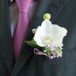 A close-up photograph captures a charcoal suit jacket and matching waistcoat worn for a special occasion in Golders Green, with the camera lingering lovingly on the lapel and the finely knotted tie. The fabric of the suit is deep charcoal, almost black, with a smooth, tightly woven texture that reflects the soft indoor light in gentle highlights, suggesting an evening celebration in a North London venue. Against this dark backdrop, a rich magenta or plum-purple tie shines with a subtle sheen, its knot sitting perfectly at the collar of a pale grey dress shirt with a barely-there woven pattern. Pinned neatly to the left lapel is an exquisite boutonniere centred on a pristine white Phalaenopsis orchid, its broad, velvety petals curving outward like sculpted porcelain. At the heart of the orchid, warm yellow and soft orange markings glow, drawing the eye inward. Around this focal bloom, delicate sprigs of lavender or pale purple blossoms and tiny white clouds of baby's breath create a dreamy, romantic halo, supported by small, glossy green leaves. A single, pale green orchid bud rises just above the main flower, promising new life and echoing the hopeful mood of a wedding morning. The stems are neatly wrapped and finished with a slender satin ribbon in soft lavender or mauve, tied in a precise bow that sits just above the breast pocket. The entire composition feels quietly refined and thoughtfully coordinated, perfect for a groom or groomsman stepping out onto the streets around Golders Green station, ready for a wedding, black-tie event, or elegant celebration nearby.