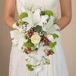 Against the soft, neutral backdrop of her lace wedding gown, a bride stands in the heart of Golders Green, holding an extraordinary cascading bridal bouquet that radiates artistry and celebration. Dominated by glorious white oriental lilies with large, outward-flaring petals delicately dusted with subtle russet spots, the bouquet immediately commands attention. Central to the arrangement, a tightly curled creamy rose offers a whisper of tradition and gentle sophistication. Vibrant lime-green cushion mums-or similar pom-pom flowers-punctuate the design with cheerful vivacity, while glossy red Hypericum berries scattered throughout evoke the spirited energy of local community events near Golders Green Road. Wispy clouds of white baby's breath infuse the bouquet with a dreamy lightness, their tiny blossoms mingling with fine grass-like greens that add natural movement and depth. The pièce de résistance is the graceful cascade of snowy Phalaenopsis orchid blooms that trail elegantly downward, lending the bouquet its signature teardrop silhouette. Bathed in soft, diffused daylight, every nuance-from the velvet sheen of lilies and the playful foam of gypsophila to the natural vibrancy of the berries-comes alive, echoing the freshness and bespoke art of local florist craftsmanship. This bouquet, meticulously arranged for a wedding in Golders Green, is the epitome of botanical romance and would inspire any bride seeking a sophisticated floral accent filled with local character and enduring charm.