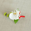 Laid gently on a pale, textured background that looks like finely woven linen or burlap, this carefully crafted boutonnière has the quiet poise of something prepared in the calm moments before a big celebration in Golders Green. The central focus is a single, immaculate white orchid, its large, silky petals unfolding in wide, graceful arcs. The petals catch the light in a way that reveals their subtle thickness and softness, while the throat of the bloom flashes with a delicate blend of yellow tones and tiny orange speckles, lending warmth and depth to the flower's otherwise cool, serene presence. Framing the orchid are soft sprays of white baby's breath, each tiny blossom like a dot of foam on the edge of a wave, offering intricate texture and a sense of airy romance. To one side sit two compact, vivid green buds that resemble button chrysanthemums, their slightly spiky, tufted forms providing both contrast and a hint of playful modernity. A single, glossy deep green leaf, reminiscent of Ruscus foliage, extends discreetly from underneath, anchoring the design with a grounded, botanical feel. The stem is tightly wrapped in a bright red satin ribbon, the material gleaming gently and gathered into a small, perfectly formed bow positioned to the right, giving the piece a refined yet spirited accent. This combination of white, green, and red feels equally at home on a groom's jacket for a synagogue wedding off Golders Green Road or on the lapel of a sharp suit heading to a formal dinner near Hampstead Garden Suburb, where the wearer might notice the faint, fresh scent and the comfortable weight of a flower arranged with practiced, local expertise.