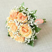 There is a quiet, understated magic to this bridal bouquet: six creamy peach roses clustered together create a soft, luminous heart, their velvety petals curling outward in concentric layers that invite touch. Encircling those roses, fine sprays of white gypsophila form a delicate, frothy veil, giving the arrangement a feeling of innocence and breathy movement as if the flowers were floating in a pale mist. The composition is punctuated by glossy, emerald-green leaves-sturdy, reflective blades that both anchor the design and heighten the warmth of the peach tones. The stems are bound in peach satin ribbon, wrapped thoughtfully for comfort, and the handle is trimmed with a neat line of small pearls whose subtle sheen catches the light when the bouquet is lifted for vows or photographs. Imagine the bouquet held on a cool morning before a ceremony at a local Golders Green venue or carried down a lane near Hampstead Garden Suburb; the subtle fragrance of rose and fresh greenery mingles with crisp outdoor air. As a florist I would note the tactile contrasts-the soft, almost velveteen rose petals against the tiny, papery gypsophila florets and the smooth, satiny ribbon-each element chosen to create a balanced, refined silhouette. The overall mood is tender and assured: an elegant piece for a bride who prefers timeless romance over ostentation, easily slipped into a pew, positioned on a cake table, or preserved as a gentle reminder of a hopeful morning.