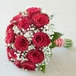 A compact, exquisitely rounded bouquet of rich red roses-each bloom fresh and velvety-commands attention against a soft, neutral canvas, their colour resonant with the warmth of local Golders Green celebrations. Crisp white clusters of baby's breath drift among the blossoms, providing an airy, textured balance to the intensity of the roses and introducing a light, celebratory spirit, much like the gentle spring breezes along Finchley Road. Hints of dark green foliage peek discreetly behind the blooms, grounding the composition with a touch of natural grace. The handle, meticulously wrapped in red and white satin ribbon and finished with a spiraling string of tiny pearls, signals both readiness and festivity-echoing the occasion's anticipation. Illuminated by soft, natural daylight, every petal, leaf, and ribbon detail is accentuated, contributing to the bouquet's timeless romantic message. This arrangement, ideal for weddings or special occasions throughout Golders Green, gracefully embodies the mingled symbolism of passion and innocence, making it a thoughtful, enduring choice for gifting and celebration across this much-loved neighbourhood.