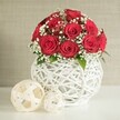 A beautifully composed floral still life captures a dome-shaped bouquet of deep, velvety red roses, styled with the poise of a wedding centrepiece that might grace a reception table in Golders Green. The roses, likely a dozen to fifteen blooms, are massed tightly together so that their open, spiralling petals touch and overlap, creating a lush, almost cushion-like surface of crimson. Threaded generously between them are fine sprays of white baby's breath, their tiny blossoms forming a delicate mist that lightens the arrangement and lends a distinctly bridal feel. Occasional glimpses of small, glossy green leaves can be seen, providing natural contrast and subtle movement. The bouquet is seated in a round, white vase with a distinctive open-weave design, made from slim strands that crisscross and loop around one another like white wicker or rattan. This airy latticework allows soft shadows to fall within the vase and gives it a handcrafted, contemporary character, the sort of piece a local florist such as Flowers Golders Green might choose for elegant wedding decor. The vase stands on a smooth, unblemished white surface, which could easily be a modern dining table set for a celebration. To the left sit two decorative woven spheres in light cream tones, one large and one smaller, echoing the material and pattern of the vase and adding gentle visual rhythm. Behind the arrangement, a softly textured, neutral-toned wall fades into blur, framing the flowers without distraction. The overall mood is serene yet romantic, perfectly suited to intimate weddings, anniversary dinners, or Valentine's Day tablescapes across NW11 and nearby Hampstead Garden Suburb.
