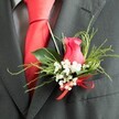 The photograph presents an intimate close-up of a classic wedding boutonniere, carefully pinned to the lapel of a charcoal grey suit jacket, evoking the polished yet romantic style often seen at ceremonies around Golders Green. The central flower is a cerise-pink rosebud, partially open so that its velvety petals fold tightly into a delicate swirl, their rich colour glowing against the dark fabric. Surrounding the rose, a halo of white gypsophila, or baby's breath, adds a lace-like softness; dozens of tiny blossoms cluster together, giving the impression of fine, airy embroidery resting on the lapel. A selection of greenery frames the flowers: slender, feathery sprigs lend a sense of light movement, while smooth, broader leaves add shine and structure, their deep green tones balancing the pink and white. At the base of the arrangement, a narrow scarlet satin ribbon is tied into a precise bow, its bright hue echoing the rich red silk tie that lies under the lapel and drawing the eye down the line of the suit. The fabric of the charcoal jacket has a subtle, finely woven texture, paired with a softly patterned grey shirt visible behind the tie, giving the entire outfit a tailored, composed feel. This buttonhole appears designed for meaningful occasions-a groom, best man, or cherished guest dressed with quiet pride, ready to step out from a home near Golders Hill Park or down Finchley Road toward an unforgettable Golders Green celebration.