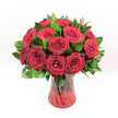 A lavish, tightly gathered bouquet of deep crimson red roses overflows from a ruby-red glass vase, creating a dramatic focal point against a clean white background. Each rose head is perfectly sculpted, the velvety petals spiralling from a tightly furled heart into generous, open whorls at the outer edge, so you can almost feel their softness with your fingertips. The rich, saturated red has the depth of fine garnet, evoking romance, passion, and a sense of classic, old-world elegance that would feel just as at home in a townhouse on Finchley Road as in a cosy flat off Golders Green Road. Nestled between the roses is a collar of dark, glossy foliage-likely Ruscus or a similar filler green-its polished leaves providing a cool, fresh contrast that makes each bloom stand out more vividly. The stems gather neatly inside a translucent red glass vase whose twisted, spiralled pattern catches and bends the light, throwing subtle highlights that echo the curves of each petal. The vase has a slightly flared rim and a sturdy base, grounding the arrangement with quiet sophistication. Bright, even lighting bathes the scene, emphasising the impeccable freshness of every rose and leaf, with not a single bruised petal or drooping stem in sight. The effect is one of pure, undiluted romance: a timeless bouquet perfect for an anniversary dinner at home near Golders Hill Park, a Valentine's surprise, or a heartfelt declaration of love. It feels like a visual love letter, composed in layers of red and green, luxurious yet sincere, ready to bring warmth and intimacy to any Golders Green living room or dining table.
