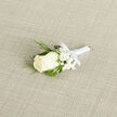 A classic wedding boutonnière lies neatly on a light, woven fabric backdrop, its understated elegance captured as though moments before being pinned to a lapel in Golders Green. At the heart of the design is a single ivory rose, its petals creamy and smooth, curling inward with a soft, romantic fullness. Around the rose gathers a gentle halo of white baby's breath, their tiny star-shaped flowers clustering in airy sprays that soften every edge and hint at new beginnings. Threaded through and around these blooms are slender ribbons of ornamental grass, deep green and slightly glossy, looping in relaxed arcs that bring an almost meadow-like freshness, as if inspired by a stroll through nearby Golders Hill Park or the leafy streets off North End Road. The stems are meticulously wrapped in white satin ribbon, its sheen catching the light just enough to signal formality without stealing the scene. The ribbon is pulled tight and secured in a discreet little bow at the base, creating a clean, polished finish that speaks of skilled, practiced hands. Resting on the calm, neutral linen, each element stands in graceful balance: the plush rose, the airy gypsophila, the structured blades of grass. You can almost imagine the subtle scent of the rose mixing with cool morning air before a ceremony at a local Golders Green synagogue or registry office. This boutonnière feels intimate and thoughtful, a small yet meaningful detail crafted to be worn close to the heart on a wedding day or special celebration in NW11.