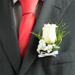 Pinned neatly to the breast pocket of a dark charcoal suit, this white rose boutonniere is shown in close-up, revealing the level of craftsmanship that a seasoned Golders Green florist would bring to a wedding morning. The rose itself is a soft ivory-white, its petals perfectly layered in a natural swirl that feels both elegant and unforced, like a quiet smile shared outside a reception venue on Golders Green Road. Nestled closely around the base of the bloom, tufts of white baby's breath create a gentle cushion, their tiny blossoms forming a fine, airy texture that softens the formality of the suit. Slender, glossy green leaves and light, feathery ferns stretch outward in smooth arcs, adding a fresh, garden-like energy that calls to mind early walks through Golders Hill Park or Hampstead Heath on a clear day. At the stem, a narrow white ribbon is tied into a discreet, perfectly centred bow, the satin catching the light in a subtle sheen. The contrast between the deep charcoal fabric and the luminous white petals heightens the boutonniere's sense of ceremony, giving the impression of a carefully chosen detail for a groom, best man, or prom date arriving in a classic car along Finchley Road. Though the image is still, you can almost sense the faint perfume of the rose and the rustle of fabric as the wearer moves. Every element-petal, leaf, ribbon, and placement-works together to express tenderness, tradition, and anticipation for the celebrations just beginning.