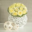 A serene, contemporary floral still life is arranged on a bright white table, capturing the eye with a rounded bouquet of light yellow roses that immediately suggests a wedding table arrangement or special celebration in Golders Green. The roses are tightly clustered into a smooth dome, each flower at full openness, revealing layers of gentle, buttery petals that create a rich yet understated texture across the surface. Threaded through the roses are fine sprigs of white baby's breath, their minuscule blossoms forming airy pockets of white that hover like tiny clouds, providing contrast and a sense of depth. Just at the base of the bouquet, a slim ring of fresh green foliage curves neatly around the arrangement, lending a crisp outline and a cool counterpoint to the warm tones above. Supporting this floral composition is a sculptural white vase, crafted from interwoven strands that cross and loop to form an open, basket-like lattice. Its rounded, modern shape feels both organic and deliberate, and the gaps between the strands let light filter through to the table below, where soft, patterned shadows appear. To the side, two pale, woven spheres made from natural-looking strips of material echo the same airy structure, as if they were small art pieces chosen to complement the flowers. The background is a softly blurred wash of neutral beige, calm and unobtrusive, like the gentle walls of a boutique venue off Finchley Road. Overall, the scene evokes a sense of quiet celebration and refined taste, the kind of centrepiece that could anchor a wedding breakfast or anniversary dinner in one of Golders Green's intimate local restaurants.
