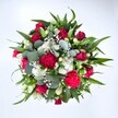 Photographed from directly above against a clean white background, this abundant round bouquet looks as if it has just been set down on a florist's bench in Golders Green. At its centre, velvety red roses in various stages of opening create a pool of rich colour, some still in tight buds, others just beginning to unfurl their layered petals in a quiet promise of romance. Around them, deep fuchsia and ruby red carnations add a ruffled, almost cloud-like texture, their fringed edges catching the light and giving the arrangement a festive, celebratory energy. Clusters of small white spray chrysanthemums and other daisy-like blooms punctuate the crimson tones with bright, crisp highlights, like little pockets of sunshine. Threaded delicately throughout, tiny white gypsophila - baby's breath - forms a fine mist between the larger flowers, softening the transitions and giving the bouquet a weightless, airy feel. Encircling the blossoms is a generous halo of greenery: strong, pointed dark green leaves, likely from lilies or alstroemeria, radiate outward to create structure, while rounded, silvery-green eucalyptus leaves nestle between petals, adding a cool, contemporary note and hinting at their fresh, herbal fragrance. The composition feels full yet balanced, with no gaps, like a carefully curated gift ready for delivery to a home near Golders Hill Park or a cosy café off Finchley Road. The mood is one of joy and heartfelt affection, perfectly suited to anniversaries, romantic gestures, or a vibrant get well soon surprise that brings a burst of colour and life indoors, even on a grey North London day.