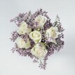 High above a small, perfectly rounded bouquet, the camera looks straight down onto six creamy white roses, each bloom tightly cupped yet softly layered, like little spirals of satin unfolding in slow motion. Their petals are a luminous ivory with the faintest blush of green at the outer sepals, giving them a fresh-from-the-cooler look that any florist in Golders Green would recognise at the start of the day. Around these central roses, a dense halo of tiny lilac and lavender limonium blossoms creates a misty, cloud-like ring, their papery petals forming a gentle haze of purple that sets the roses delicately aglow. The filler flowers are packed closely enough to feel lush, yet light and feathery in texture, the way a breeze might ripple through the borders in nearby Golders Hill Park. Everything rests on a smooth, pale off-white surface, calm and uncluttered, so the flowers remain the absolute focus. Soft, even lighting falls from above, revealing every vein in the rose petals and every tiny star-shaped lilac floret without harsh contrast, as if the bouquet is sitting by a north-facing window on a quiet London morning. The overall mood is serene and deeply comforting, a design that would suit a sympathy gesture, a thoughtful thinking of you delivery around Golders Green Road, or a gentle bridal posy. You can almost imagine this hand-tied design being carried out from a local studio, its white and lavender tones bringing a sense of peace, purity, and understated elegance to any home or bedside table nearby.
