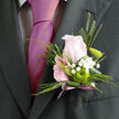 The image focuses tightly on an elegant boutonnière, the kind you might see being carefully pinned on just before a ceremony at a synagogue or town hall near Golders Green. At its centre rests a pale pink rose, its petals gently unfurling in a soft spiral, as if caught midway between bud and bloom. The colour is delicate, almost like blushed porcelain, and it gives the whole piece a romantic, almost storybook quality. Around the rose, slender pieces of foliage and a neatly folded green leaf are arranged with a clean, modern sensibility, adding structure without overwhelming the flower's natural grace. Threads of white gypsophila trail along the stem, each tiny, star-shaped bloom catching the light and giving a misty, cloud-like softness that contrasts beautifully with the rose's smooth curves. Tucked in beside the rose is a vivid green button chrysanthemum, round and textured, lending a contemporary pop of colour that feels fresh and youthful, ideal for weddings or proms across Golders Green and nearby Temple Fortune. The stems are expertly wrapped in a muted mauve ribbon, its satin surface catching a gentle sheen, with a small tail of the ribbon creating a quiet, decorative flourish beneath the blooms. The overall effect is one of calm refinement: a small, hand-crafted piece that reflects both tradition and modern styling, ready to complement a tailored suit and carry the subtle perfume of the rose through a day of smiles, photographs, and heartfelt moments.