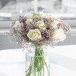 A full, rounded bouquet of creamy white roses and soft lilac statice stands gracefully in the centre of the image, arranged with the quiet confidence of an experienced Golders Green florist. Around a dozen white roses, each bloom just beginning to unfurl, reveal layers of velvety petals in an ivory shade that feels both modern and timeless. Between the roses, clouds of tiny lavender-purple statice create a misty halo, their fine, papery blossoms adding lightness and an almost feathery texture that softens the overall silhouette. The flowers are gathered in a tall, clear cylindrical glass vase, where the long green stems are visible beneath the waterline, clean and straight, hinting at freshly cut blooms prepared for same-day delivery across Golders Green and nearby Hampstead Garden Suburb. The vase rests on a smooth, bright white tabletop that echoes the purity of the roses and reflects the gentle daylight streaming in from a softly blurred background. The light appears to come from a large window, similar to those overlooking Golders Hill Park, casting a calm, luminous glow over the petals and creating faint, tender shadows. There is no clutter around the arrangement, allowing the bouquet's tranquil presence to fill the space. The mood is serene and contemplative, suited to a sympathy gesture, a quiet anniversary at home off Finchley Road, or simply a soothing floral accent in a contemporary flat. Even without scent in the image, you can almost sense a delicate rose fragrance mingling with a hint of fresh greenery, as if the bouquet were bringing a breath of peaceful garden air into a still, sunlit room.