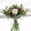 A serene bouquet of white roses, lavender alstroemeria, and airy baby's breath stands poised in a clear cylindrical vase, bathed in soft, diffused light reminiscent of a calm morning in Golders Green. The roses form the heart of the composition, their generous blooms fully unfurled, each petal thick, smooth, and gently curling, creating rounded rosettes of creamy white that feel both pure and reassuring. Around these central flowers, sprays of gypsophila scatter tiny white blossoms, so fine and numerous that they appear like a delicate mist, softening the lines of the arrangement and giving it a tender, almost whispering presence. Tucked between the roses are elegant alstroemeria stems, some still in slender bud, others just opening to reveal petals washed in lilac, pale purple, and muted cream, with subtle stripes that add understated character without disturbing the overall calm. Encasing the base of the floral heads, broad, polished green leaves are wrapped and overlapped with care, creating a structured collar that holds the rounded form and hints at the florist's practiced hands, perhaps working in a studio just off Golders Green Road. Finer, upright leaves interlace through the blooms, lending height and a breath of natural movement, like foliage stirred by the wind above nearby Hampstead Garden Suburb. Through the clear glass of the vase, the straight green stems are seen in crisp detail, resting in fresh water and gathered neatly with a slim green tie just above the waterline. The background is bright and uncluttered, drawing all attention to the flowers themselves. Together, these elements evoke a feeling of quiet reflection and gentle strength, making this bouquet fitting for condolence delivery or for placing in a tranquil corner during a time of mourning.