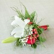 On a soft, linen-textured beige fabric that hints at the cozy charm of Golders Green homes, this beautifully arranged floral accessory radiates both grace and festivity. Dominating the design is a stately white lily, each petal gracefully curling outward and flecked with subtle reddish tones, basking in soft, natural light that accentuates its pure radiance. Alongside, a rich red rose opens in luxurious fullness, its petals lush and deeply hued. Glossy red berries-hypericum-twinkle playfully within the arrangement, adding a festive flourish that conjures holiday markets along Finchley Road. Wisps of delicate white baby's breath create gentle, airy halos, complimented by a medley of leafy greens ranging from feathery ferns to broader, lustrous leaves. To the left, a slim, green lily bud hints at hopeful new blooms and future celebrations. The stems are thoughtfully gathered and wrapped in a vibrant red satin ribbon-a detail that ties together the entire palette with artisanal flair. Brimming with emotion and artistry, this compact bouquet embodies Golders Green's vibrant spirit, suited for weddings, school proms, or heartfelt gifts, and expertly crafted for the joyful moments that bring the local community together.