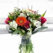 This vibrant bouquet captures the spirit of a radiant Golders Green morning, expertly crafted by local florists and presented in a clear glass vase that allows every verdant stem to shine. At its centre blazes a joyful orange Gerbera daisy, its petals a vibrant halo around a striking green heart, evoking warmth and energy reminiscent of the lively atmosphere along Finchley Road or the community gatherings at Golders Green's cafés. Encircling the Gerbera, velvety roses in passionate shades of magenta and crimson offer romance and sophistication, their blossoms in various stages of bloom suggesting hope and renewal. Among these, creamy white alstroemerias curve gracefully outward, their striped petals and speckled throats creating a fresh, uplifting counterpoint, interlaced with tiny daisy chrysanthemums that bring a whimsical, airy charm. Cream and buttery yellow roses, along with the gentle hush of pale lavender florets, layer in softness and depth, completing the bouquet's symphony of colour. Glossy, broad green leaves sweep outward from the base, embracing the arrangement with organic movement, while slender, spiky foliage adds a sculptural touch. The local daylight spilling through a sunlit window bathes the ensemble, making every colour glow with vivid clarity. Perfect for moments of congratulations, a birthday surprise, or simply to brighten a day, this bouquet feels as if it holds a piece of Golders Green's welcoming spirit-fresh, heartfelt, and designed to share happiness with those you cherish.