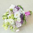 A visually striking hand-tied bridal bouquet rests diagonally against a soft cream linen background, softly bathed in natural, even light that enhances each gentle hue. Front and center, large white Lisianthus blossoms unfurl their ruffled petals, accented by tender lemon-yellow hearts, surrounded by a festival of pale green buds and wispy lavender-pink stock. Bold deep purple allium florets explode like city fireworks, their dramatic texture providing lively contrast with the softness of the Lisianthus. The bouquet's bright green foliage, looping and arching across the composition, recalls walks through Golders Green's well-kept parks in early spring. Tightly gathered stems are bound in a fuchsia pink satin ribbon, woven with lustrous white pearls-a statement of modern romance and timeless style. Whether for a wedding in one of Golders Green's elegant venues or a celebratory gathering with friends, this bouquet encapsulates freshness, grace, and the understated luxury of expert floral design, making it a perfect visual for stories on wedding trends and local floral artistry.