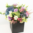 A luxurious, garden-style bouquet sits proudly in a sleek matte black square box, photographed against a crisp white background so every colour appears luminous and true. The arrangement is densely packed and softly rounded, its heart formed by generous clusters of hydrangeas in deep periwinkle, dusty blue, and hints of mauve, their plush petals layered like little clouds. Threaded through this cool, velvety base are graceful lavender and soft blush-pink roses, some tightly furled, others just beginning to unfurl in a classic spiral, adding a gentle romantic note. Creamy white lisianthus, with their slightly ruffled, trumpet-shaped blooms and delicate buds on slender stems, rise lightly above the hydrangea mounds, bringing a fresh, airy brightness that catches the eye much like morning light over Golders Green Station. Here and there, small tufts of vivid lime-green textured blooms, reminiscent of Green Trick dianthus, introduce playful, mossy spheres that contrast beautifully with the smoother rose petals. Sprays of fine, feathery pink astilbe and other petite filler flowers create wisps of movement, as though a breeze from nearby Golders Hill Park had just brushed past. Varied greenery, from bright new shoots to deeper green leaves, fills the gaps and frames the richer blossoms, making the composition feel both abundant and carefully balanced. The bold black box acts as a modern pedestal, giving the design a contemporary, London-chic elegance that would suit a stylish flat near Finchley Road as easily as a reception desk on Golders Green Road. The overall mood is polished yet heartfelt, perfect for thoughtful congratulations, an elegant birthday surprise, or a tender gesture of comfort delivered locally the same day across NW11.