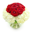 A refined and romantic floral design is displayed in a round, clear glass vase, set against a bright white background that makes the colours appear almost luminous. The eye is immediately drawn to the domed cluster of deep red roses at the top, roughly ten blooms nestled tightly together so that their velvety petals touch and intertwine. Some roses are fully open, showing off intricate spirals of crimson, while others are just beginning to unfurl, giving a sense of movement and life. Beneath this lush crown of roses sits a voluminous mass of creamy white hydrangeas, carefully arranged to form a smooth, spherical base. The hydrangea florets are small and abundant, creating a soft, cushiony texture that supports the roses above and provides a beautiful visual contrast. The transition from intense red to gentle white feels deliberate and harmonious, like a visual representation of passion resting on a foundation of calm, enduring affection. The clear water inside the bowl-shaped vase reveals the stems gathered neatly together, their green tones adding a subtle hint of freshness and grounding the design. The neutral background keeps all the focus on the flowers, with no distractions from their luxurious textures and rich hues. From a local Golders Green perspective, this arrangement feels perfect for an intimate anniversary celebration-perhaps delivered to a home near Golders Hill Park or to a favourite restaurant table as a surprise gesture. It is polished yet deeply heartfelt, ideal for saying, Our love is strong, steady, and still beautifully alive, in a way that feels both classic and contemporary.