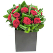 In this elegantly modern composition, deep crimson roses form the quiet, dramatic centre of the design, each bloom unfurling velvety petals in tight, generous spirals that catch the eye. Broad, glossy green foliage has been carefully curled and intertwined around the roses, the leaves forming a rhythmic pattern of overlapping arcs that add motion and sculptural depth. The arrangement sits in a matte black cube - a square, box-like vase whose flat planes ground the bouquet and provide a contemporary contrast to the soft organic forms above. Bright, clear studio lighting picks out the velvet sheen on each petal and the glossy highlights along the curled leaves, giving the whole piece a refined, almost urban polish. Close to the blooms you can imagine a subtle aroma of fresh rose rounded by crisp green notes from the foliage, a scent that reads as clean and uplifting rather than heavy. Texturally there is a delightful dialogue between the silk-like softness of the petals and the firmer, almost waxy texture of the leaves; every crease and fold has been placed with intention. As a florist I see this as a design suited to modern spaces in Golders Green - a striking centrepiece for a contemporary dining table or a thoughtful gift to mark a romantic occasion, an anniversary, or a meaningful thank-you. The overall feeling is composed yet emotive: bold in colour, restrained in form, and quietly unforgettable, a bouquet that speaks with confident simplicity while retaining a hand-crafted warmth.