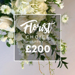 An opulent white-and-cream spray fills the frame with luxuriant blooms and garden-fresh greens, as if harvested from a quiet corner of Golders Green. At the core are large creamy white roses, fully opened and silken to the touch, paired with generous clusters of pale hydrangeas that lend volume and softness. A cascade of white sweet peas or similar slender blooms threads down the length of the arrangement, while Nigella flowers peek through near the top, their green calyxes and feathery foliage lending architectural bite. The greenery is varied and lush: broad deep-green leaves, light variegation for contrast, and fine curling fronds that drift below the main cluster. The piece sits within a clean white square frame, the overlay text proclaiming Florist Choice in a graceful script, followed by SPRAY in crisp uppercase letters and a bold ?200 at the base. The soft, neutral backdrop enhances the pristine whites and greens, signaling a premium bespoke design available for same-day delivery across Golders Green, Temple Fortune, and the surrounding NW11 area. This image speaks of refined celebration, quiet remembrance, and the skilled hands of a local florist crafting beauty for birthdays, anniversaries, or sympathy tributes, all proudly offered by Flowers Golders Green.