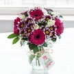 A vibrant mixed bouquet of magenta gerbera daisies, purple-tipped white chrysanthemums, creamy white roses, and airy sprigs of baby's breath stands proudly in a clear, textured glass vase, set against a softly blurred white backdrop that feels like daylight filtering through a Golders Green window. The gerberas, in deep fuchsia and rich pink tones with dark central disks, form bold, cheerful focal points, while the chrysanthemums display intricate petals edged in velvety purple that melt into crisp white around bright yellow centres. Nestled between these statement blooms, clusters of small white spray roses, some fully open and others still in tight bud, lend a gentle, romantic softness. Fine clouds of white gypsophila weave delicately through the arrangement, adding a light, almost sparkling texture that contrasts with the lush, glossy green foliage at the base. The stems are clearly visible through the squat, jar-style vase, which is cinched at the neck with a simple loop of rustic twine tied into a bow. A small, rectangular cream tag hangs from the twine, printed with a pink rose motif and elegant green lettering, hinting at a personal message from a local sender. The whole design is clearly the work of an experienced florist, evoking the feeling of a thoughtful surprise delivered to a quiet home just off Finchley Road or near Golders Hill Park. Bathed in gentle natural light that makes each petal glow, the bouquet radiates joy and celebration, ideal for marking birthdays, thank-you gestures, or spontaneous thinking of you moments in and around Golders Green.
